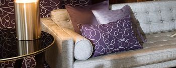 Elegantes Wohnzimmerdetail mit einem grauen gepolsterten Sofa mit lila gemusterten Kissen, einem Beistelltisch aus Glas und einer modernen Lampe mit Akzenten von Produkte. Raumausstattung Merget | Goethestraße 43 | 63814 Mainaschaff
