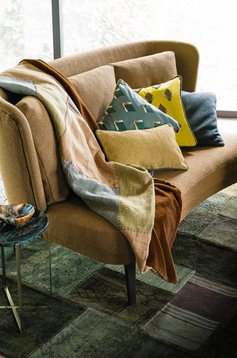Ein gemütlicher Wohnraum mit einer braunen Couch, bedeckt mit bunten Decken und Kissen, neben einem Fenster mit Gardinen und Grün draußen. Raumausstattung Merget | Goethestraße 43 | 63814 Mainaschaff